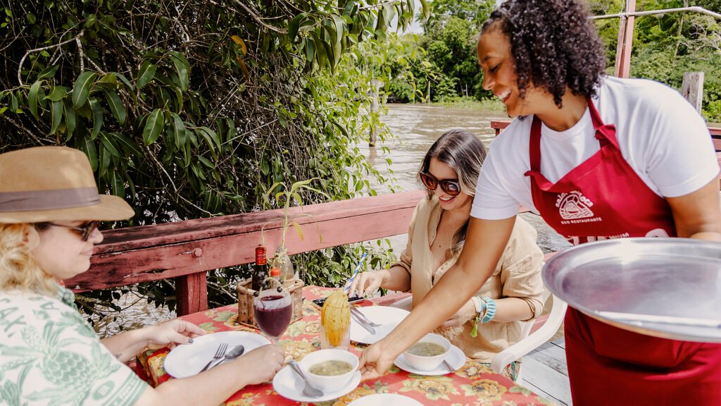 Imagem de uma atendente do restaurante Saldosa Maloca, na ilha do Combú, servindo comida para um casal sentado na mesa ao lado do rio.